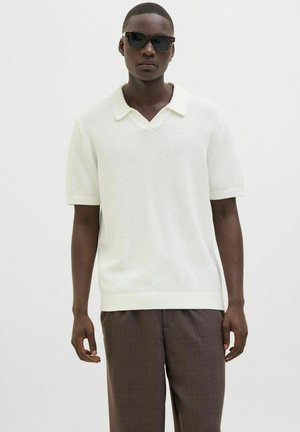 Polo en tricot blanc à manches courtes avec un col en V et un col contrastant. Le modèle porte un pantalon à carreaux marron et des lunettes de soleil foncées.