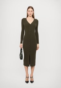 Rochie ribbed de culoare verde măslin, cu mâneci lungi, decolteu în V, brăcită pe față cu butoane și cu o fanta de lungime medie. Modelul ține o geantă neagră.