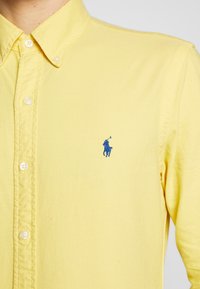 Żółta bawełniana koszula zapinana na guziki, z niebieskim haftowanym logo na lewej piersi oraz klasycznym kołnierzykiem na guziki.