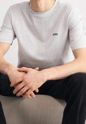 Personne portant une chemise à manches courtes gris clair avec le logo "BOSS" sur la poitrine, assise les mains jointes sur un pantalon noir, sur un siège texturé.