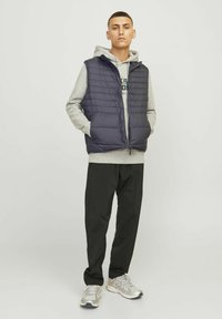 Marineblauwe puffer vest met een gewatteerd ontwerp, ritsluiting en zijzakken, gedragen over een grijze hoodie, gecombineerd met zwarte broek en zilveren sneakers.