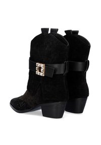 Botines de ante negro con una correa de cuero negro que presenta una hebilla dorada y strass. La puntera está adornada con tachuelas negras y un tacón ancho.