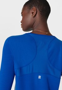 Blauwe sporttop met lange mouwen, voorzien van een textuurpaneelontwerp, gladde stof en een kenmerkend logo op de rug dicht bij de halslijn.