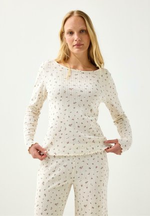 Donna con lunghi capelli biondi indossa un top bianco a maglia a costine e pantaloni con piccola stampa floreale e orli effetto lattuga su sfondo semplice.