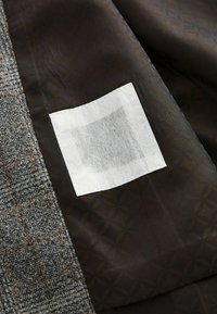 Brązowa tkanina w geometryczny wzór o gładkiej fakturze, obok szarego materiału w kratę. Z boku przyszyta jest biała metka.