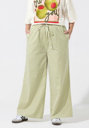 Person iført vidtbenede grønne og hvide lodrette stribede bukser med snøre i taljen, hvide sneakers og en t-shirt med olivengrafik.