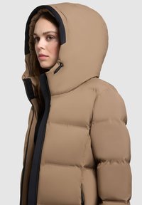 Kurtka puchowa w jasnobrązowym kolorze z dużym kapturem. Posiada suwak z przodu, regulację na troczki oraz pikowaną fakturę. Czarne akcenty wewnątrz.