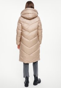 Lange beige pufferjas met een chevronpatroon, een grote capuchon en een gewatteerde textuur, gedragen over grijze broek en zwarte enkellaarzen.