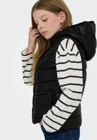 Crna pufasta prsluk s kapuljačom, s horizontalnim kvadratićima. Nošen preko bijele i smeđe prugaste majice dugih rukava.