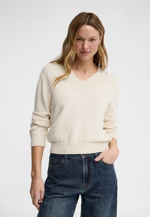 Kvinde med langt blondt hår iført en cremefarvet V-hals sweater og blå jeans, stående med den ene hånd i lommen mod en ensfarvet baggrund.