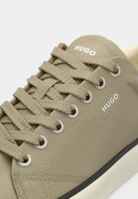 Μπεζ καμβάς αθλητικό παπούτσι με υφή στην επιφάνεια, στρογγυλεμένη μύτη και λευκές οπές για τα κορδόνια. Διαθέτει χοντρά κορδόνια και μικρό λευκό λογότυπο στο πλάι.