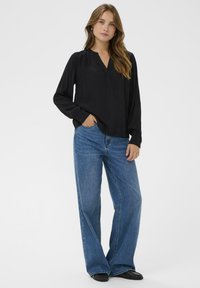 Sort langærmet bluse med V-hals og indsnit ved skuldrene, parret med brede blå jeans og sorte flade sko.