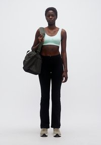Γυναίκα που φοράει ανοιχτό μπλε αθλητικό σουτιέν και μαύρο παντελόνι, κρατώντας μια σκούρη πράσινη τσάντα γυμναστηρίου στον ώμο, στέκεται μπροστά σε απλό φόντο.
