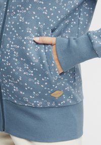 Blå zip-up hoodie med ljusrosa och blå prickmönster, ribbade cuffs och fåll, med en logopatch nära fickan.