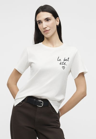 Γυναίκα με μακριά σκούρα μαλλιά που φοράει λευκή μπλούζα με το κείμενο «le bel été» και καρδιά, σκούρο παντελόνι και μαύρη ζώνη, σε απλό φόντο.