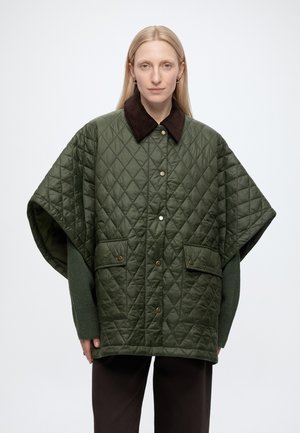 Femme blonde portant une cape matelassée oversize vert olive avec un col marron foncé et des boutons-pression à l'avant, debout sur un fond uni.