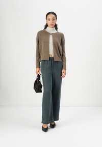 Brown knit cardigan over a striped turtleneck, paired with relaxed dark green trousers and a small black handbag. Flats complete the outfit.