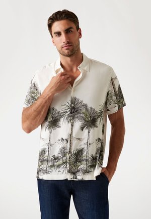 Camisa de botones de manga corta en color crema con estampado de palmeras y montañas. Tejido ligero, corte relajado y textura sutil. Combinada con vaqueros oscuros.