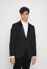 Blazer nero con lapel in raso, due tasche frontali e chiusura a un bottone, indossato sopra una camicia bianca.