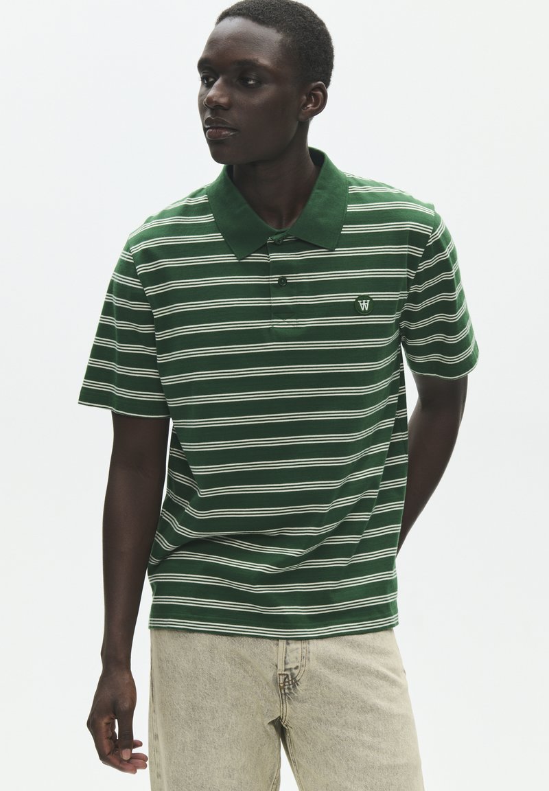 Modèle masculin portant un polo à rayures vertes et blanches et un pantalon beige clair, debout avec une main derrière le dos.