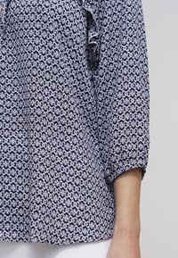 Blouse à motifs présentant un design floral bleu marine et blanc, manches longues avec des poignets retroussés, et une texture de tissu douce et légère.