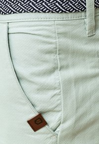 Pantalones cortos verde claro hechos de tela texturizada, con un bolsillo lateral y una etiqueta de logo marrón. Cintura con patrón en blanco y negro visible.