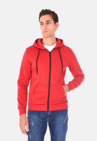 Felpa rossa con zip e tasca frontale, cappuccio regolabile con lacci e zip nera. Indossata sopra una camicia bianca e abbinata a jeans blu.