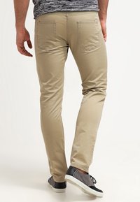 Homme portant un pantalon slim beige et des baskets grises, se tenant avec un pied légèrement levé, faisant face à l'opposée de la caméra.