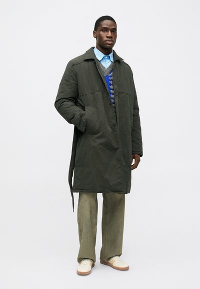 Homme debout portant un long manteau vert foncé, une chemise bleue, un pull à motif, un pantalon vert ample et des baskets blanches devant un fond unicolore.