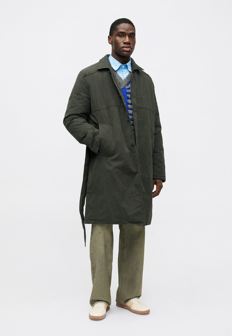 Bărbat în picioare, îmbrăcat într-o haină lungă de culoare verde închis, cămașă albastră, pulover cu model, pantaloni verzi lejeri și adidași albi, pe un fundal simplu.