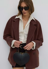 Hnědý oversize blazer z vlněné směsi, bílá pruhovaná košile, černá kabelka ve tvaru srdce se zlatými akcenty a černé sluneční brýle.