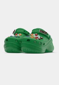 Green clogs made from rubber-like material feature a chunky sole, ventilation holes, and colorful M&M’s decorations on the upper part.