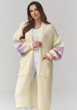 Kobieta z długimi brązowymi włosami nosi kremowy długi sweter z dzianiny z pastelowymi paskami na balonowych rękawach, biały top i białe spodnie, stoi na tle szarego tła.