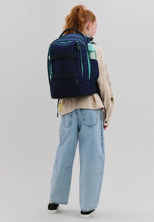 Junge Person mit roten Haaren, trägt eine beige Jacke und weite blaue Jeans, hat einen dunkelblauen Rucksack mit einer Wasserflasche in der Seitentasche.