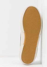 Braune Sneaker-Außensohle mit genoppter Textur, ausgestattet mit einem weißen Gummirand und einer flexiblen, hellbraunen Gummisohle für Griffigkeit und Langlebigkeit.