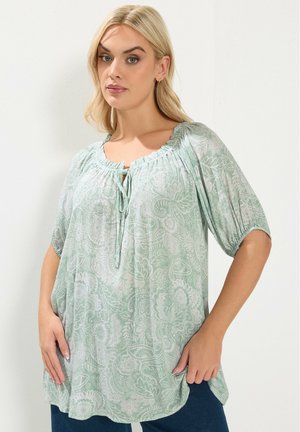 Kobieta o blond włosach, ubrana w jasnozieloną bluzkę w kwiatowy wzór z bufkami na rękawach oraz niebieskie spodnie, stojąca z rękami blisko bioder.