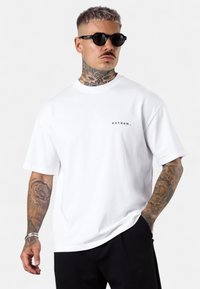 Weißes Baumwoll-T-Shirt mit kurzen Ärmeln, auf der linken Brust mit einem kleinen schwarzen Textlogo "esteem." versehen. Das Model trägt schwarze Sonnenbrillen.
