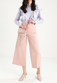 Blau-weiß gestreifte Bluse mit Volants, blassrosa weite Hosen und eine hellrosa Umhängetasche mit Fransen; beigefarbene Mules vervollständigen das Outfit.