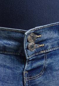 Jeans de denim azul con una cintura alta, que presentan dos botones plateados, costuras en contraste y una sutil textura de la tela visible.