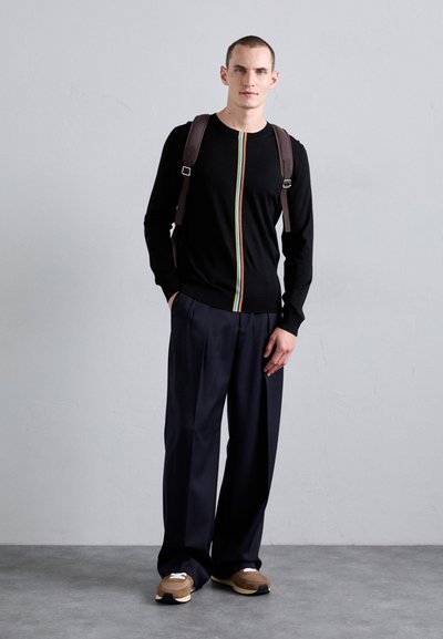 Svart langermet genser med en sentral regnbue-stripe, kombinert med løse marineblå bukser og brune sneakers med hvite detaljer.