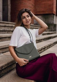 Šedá kožená kabelka s klopou a zlatým logem, kterou nese osoba v bílém tričku a burgundské sukni, sedící na kamenných schodech.