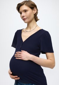 Top premamá azul marino con mangas cortas abullonadas, escote en V y cierre frontal con cremallera. Tejido acanalado ajustado sobre la barriga redondeada, combinado con denim.