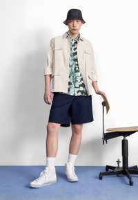 Homme portant un chapeau seau noir, une veste beige, une chemise à motifs verts, un short bleu marine, des chaussettes blanches et des baskets blanches, se tenant à côté d'une chaise pivotante en bois.