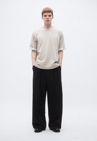 Beiges T-Shirt mit kurzen Ärmeln und einem Karomuster an den Bündchen, kombiniert mit locker sitzenden schwarzen Hosen und schwarzen Schuhen. Sauberes Design und minimalistischer Look.