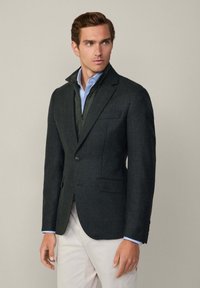 Mörkgrön texturerad blazer med två framfickor, enknappsstängning, lagd över en ljusblå skjorta. Avslappnad elegans med skräddarsydd passform.