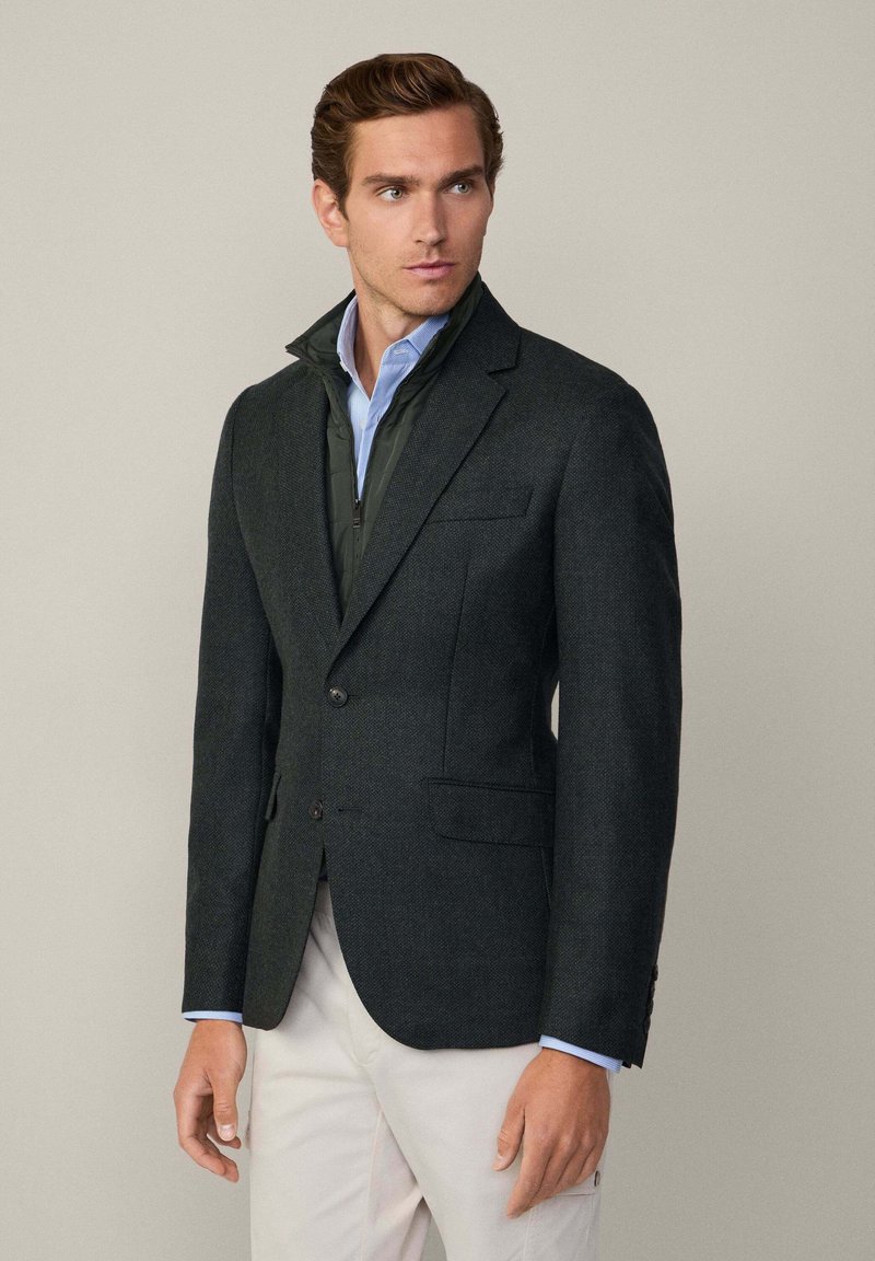 Mörkgrön texturerad blazer med två framfickor, enknappsstängning, lagd över en ljusblå skjorta. Avslappnad elegans med skräddarsydd passform.