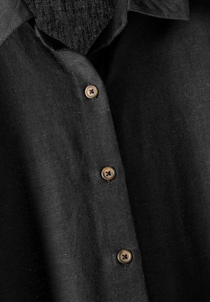 Chemise noire à boutons, en tissu texturé, avec un col pointu, cinq boutons marron et un léger brillant.