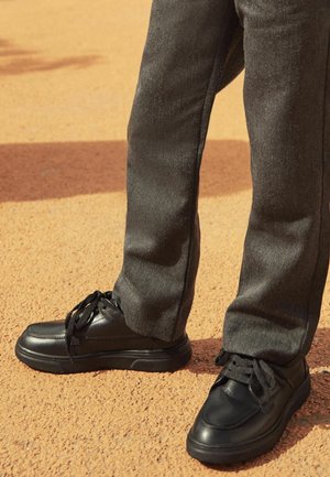 Baskets en cuir noir avec surface texturée, fermeture à lacets et semelle en caoutchouc épaisse, associées à un pantalon gris ajusté.