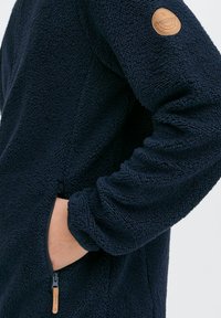 Marineblaue Fleecejacke mit strukturierten Oberflächen, einer Reißverschlusstasche und einem Leder-Logopatch am linken Ärmel. Weiches und warmes Material.