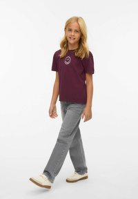 T-shirt bordeaux avec un graphique de smiley blanc, associé à un jean droit gris clair et des baskets crémeuses, marchant sur un fond neutre.